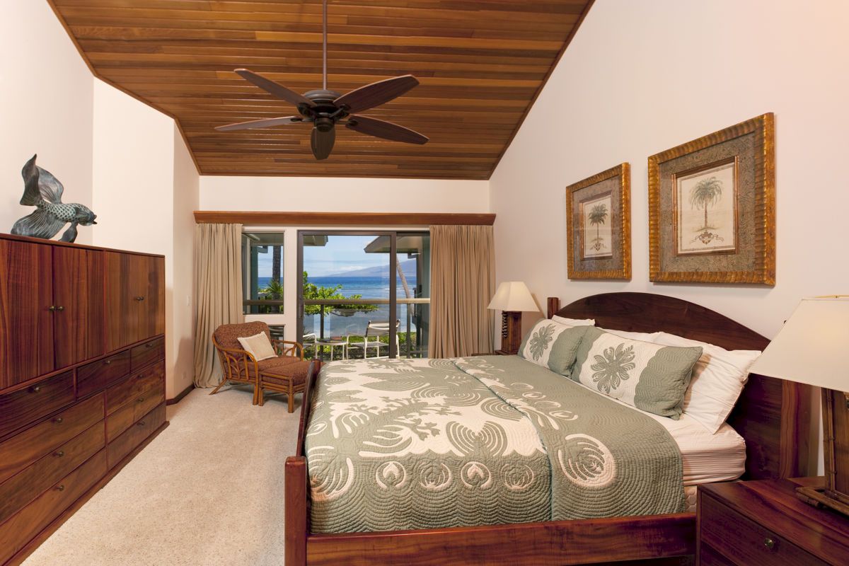 A cozy bedroom with a large bed, wooden furniture, ceiling fan, artwork on walls, and window view of the ocean and outdoor seating.