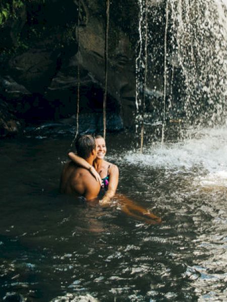 A person standing in the water near a waterfall, surrounded by lush greenery with some pink flowers in the foreground.