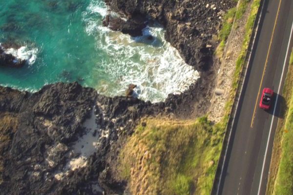 Aerial view of a coastal road with a red car driving, alongside a rocky shoreline with clear blue water and waves, bordered by greenery.