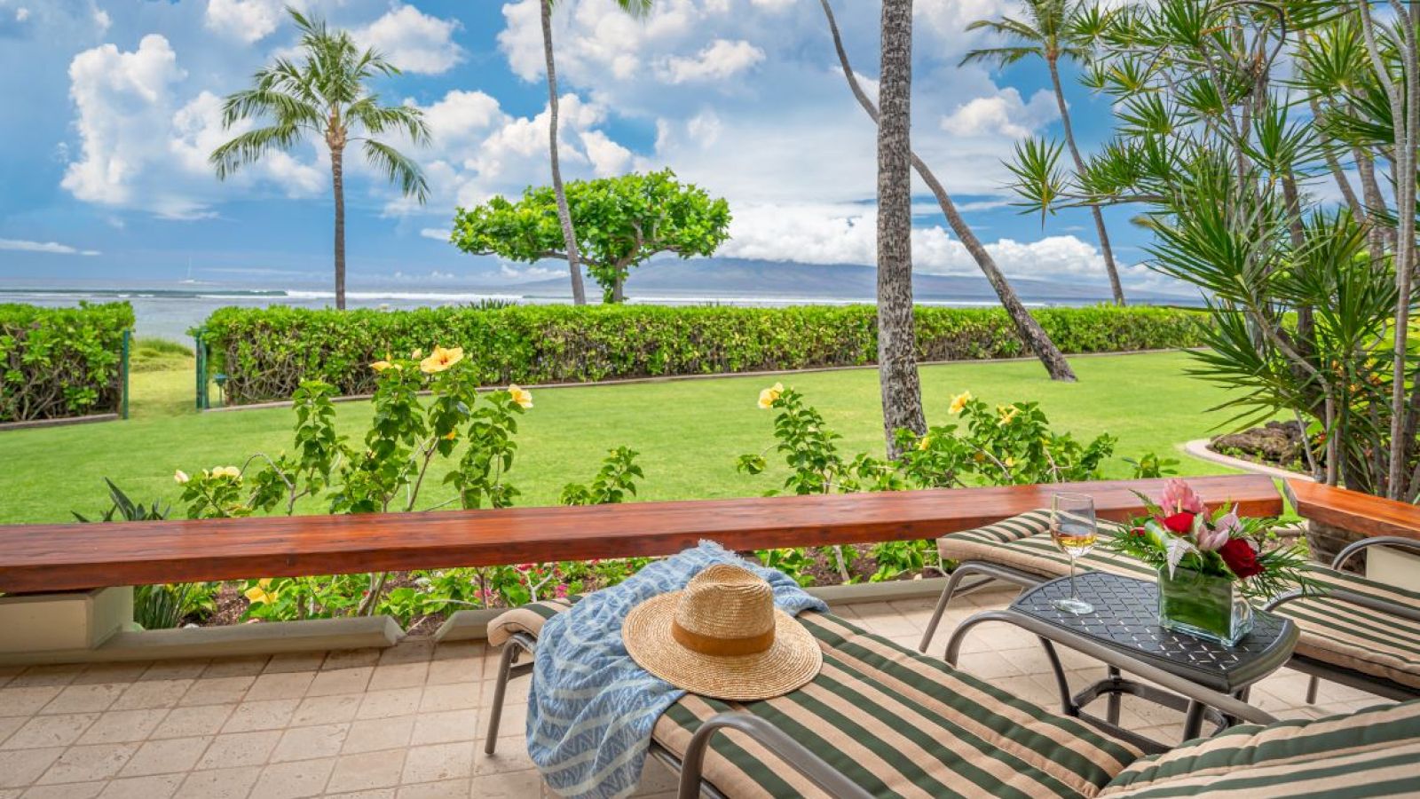 The image shows a tropical patio with lounge chairs, a sunhat, flowers on a table, lush greenery, palm trees, and a view of the ocean under a blue sky.