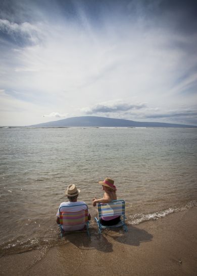 Two people sitting on beach chairs facing the ocean, with a mountain visible in the distance. The sky is partly cloudy.