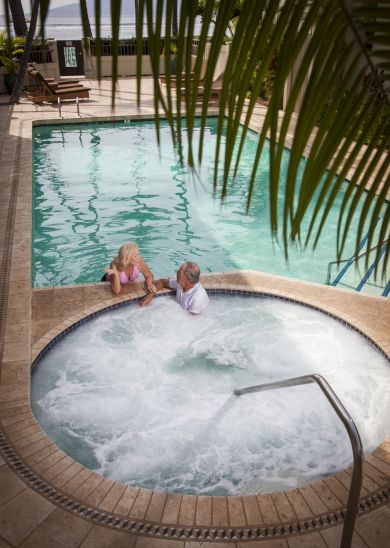 Two individuals are relaxing in a hot tub next to a pool, with deck chairs in the background and palm fronds partially obstructing the view.