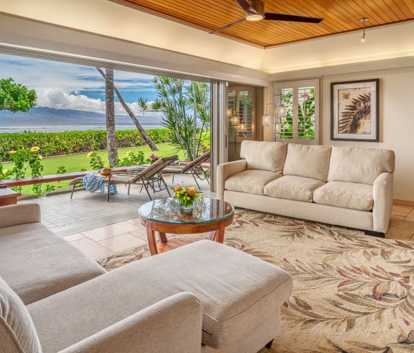 A cozy living room with two sofas, a glass coffee table, and a view of a lush outdoor area with loungers facing the ocean.