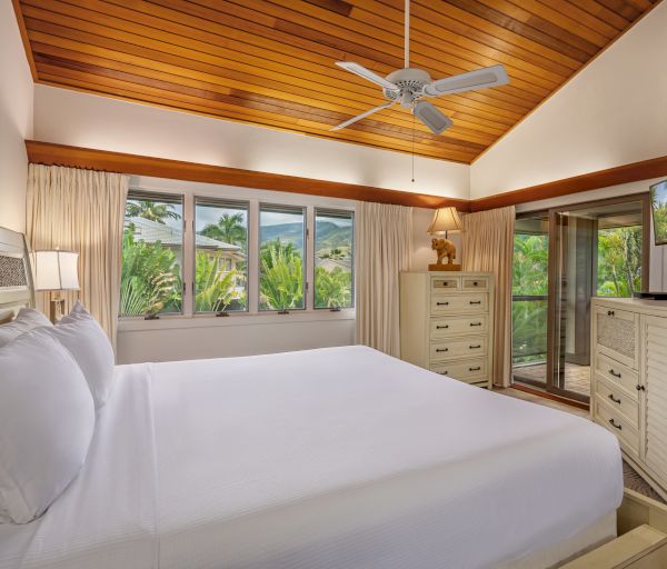 A bright bedroom with wooden ceiling, white bed, dresser, TV, ceiling fan, and large windows showing a tropical garden view, ending the sentence.