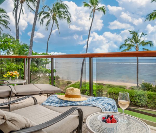 Beachfront balcony with lounge chairs, a straw hat, a drink, and fruit on a table, overlooking palm trees and the ocean on a sunny day.