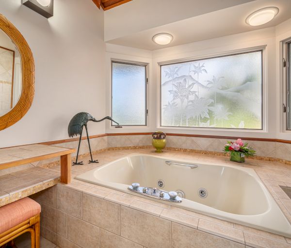 A bathroom with a large bathtub, frosted windows, a wooden vanity with a round mirror, a decorative bird figurine, and a vase with flowers.