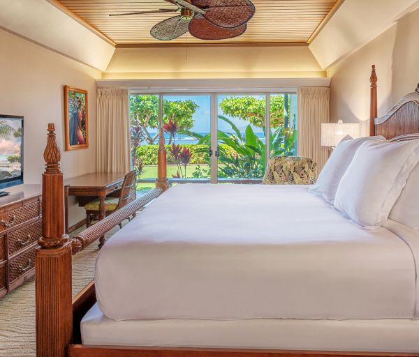 A cozy bedroom features a large bed, dresser with a TV, desk by the window, tropical artwork, ceiling fan, and a view of lush greenery outside.