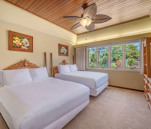 This image shows a cozy bedroom with two double beds, wooden ceiling, ceiling fan, large windows, floral paintings, and a bright, inviting atmosphere.