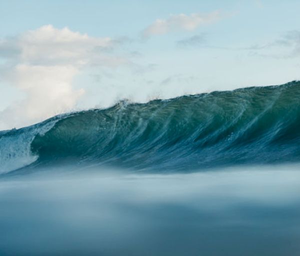 The image shows a close-up of an ocean wave about to crest, with a partly cloudy sky in the background.