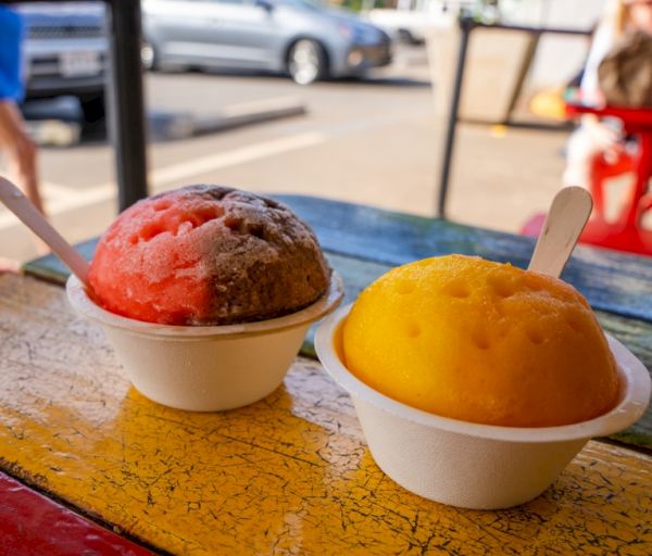 Two cups of shaved ice, one red and brown, the other yellow, on a colorful wooden table outdoors with parked cars in the background.