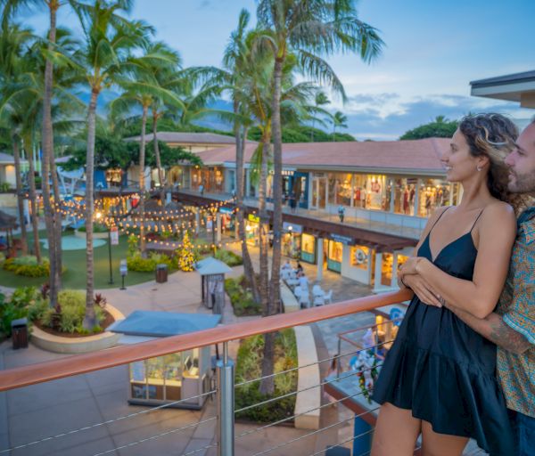 A couple stands on a balcony overlooking a beautifully lit outdoor shopping center with palm trees and a festive ambiance ending the sentence.