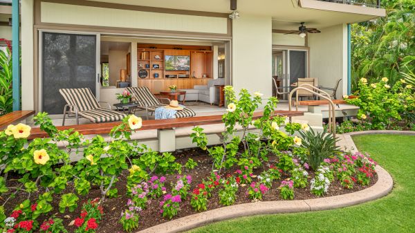 A cozy outdoor patio with lounge chairs, vibrant flowers in a garden bed, and an open view into a modern living room with wooden furniture.