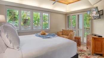 A cozy bedroom with a large bed, a hat and scarf on the bed, tropical-themed decor, wooden furniture, a TV, and windows with shutters.