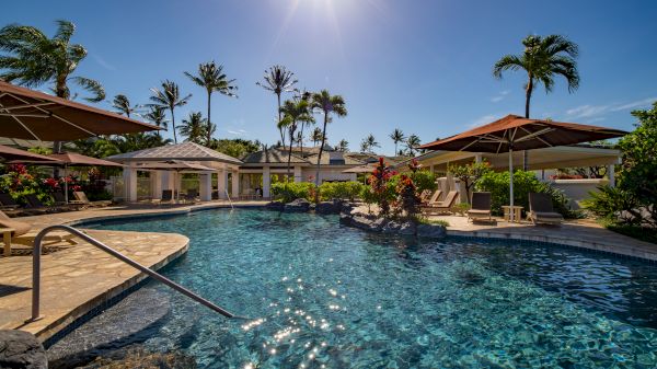 A luxurious outdoor pool area with parasols, lounge chairs, and lush palm trees under a bright, sunny sky.