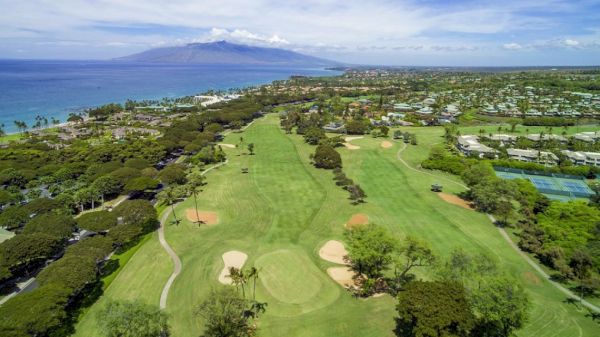 An aerial view of a scenic golf course with sand traps, surrounded by trees and houses, near an ocean with a mountainous island in the distance.