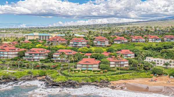 This image shows a coastal community with red-roofed buildings, lush greenery, and a sandy shoreline meeting the ocean waves, under a partly cloudy sky.