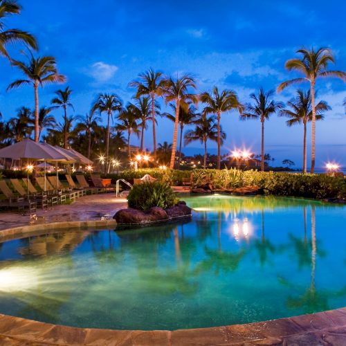 A beautifully lit swimming pool surrounded by palm trees and lounge chairs at dusk, with a scenic backdrop of the ocean and a clear blue sky.