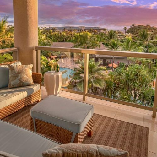 A cozy balcony setting with cushioned seating overlooking a tropical resort landscape, featuring palm trees and a pool under a colorful sunset sky.