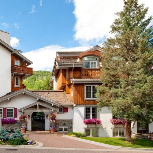 A charming alpine-style building surrounded by greenery, featuring wooden accents, balconies, colorful flowers, and a welcoming arched entrance.