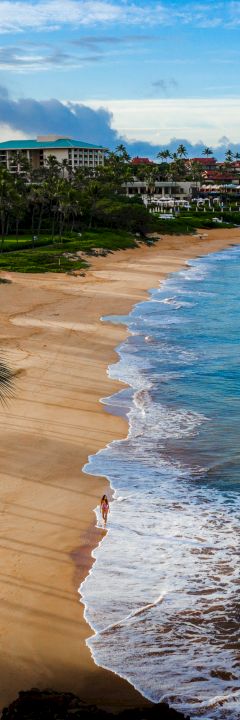 A beautiful beach scene with golden sand, gentle waves, a solitary person walking along the shoreline, and buildings in the background.