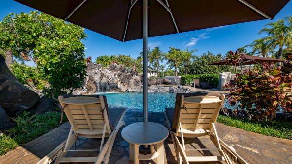 Two lounge chairs under an umbrella by the pool with a waterfall, surrounded by lush greenery and palm trees under a clear blue sky.