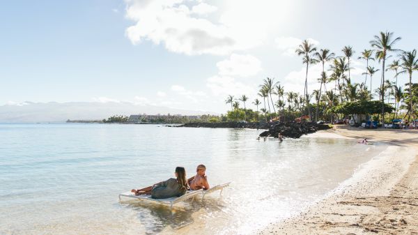 Two people relax on a lounge chair in shallow, clear water by a sandy beach lined with palm trees, under a bright blue sky, with gentle waves.