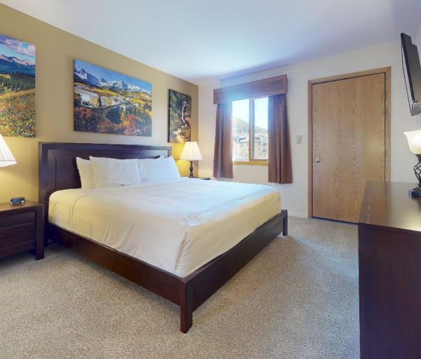 A cozy bedroom with a large bed, bedside tables, lamps, a dresser, wall-mounted TV, and nature-themed wall art. The door is closed.