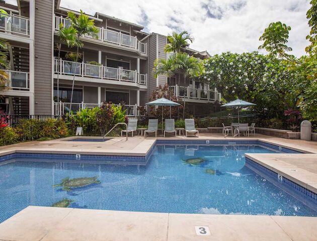 An outdoor pool area with surrounding lounge chairs, umbrellas, and a multi-story building in the background, complemented by lush greenery ending the sentence.