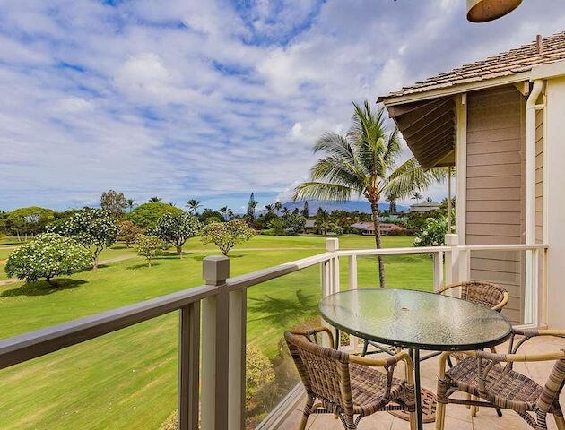 A balcony with a glass table and wicker chairs overlooks a lush golf course with palm trees under a partly cloudy sky.