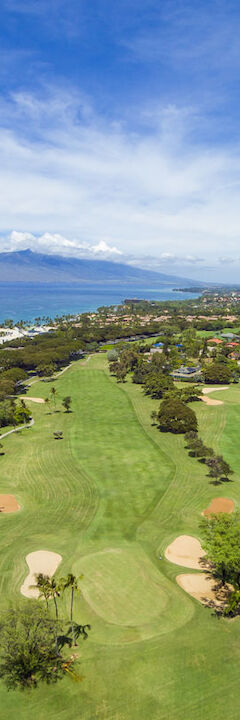 Aerial view of a lush golf course near the ocean, with mountains in the distance and a residential area to the right.