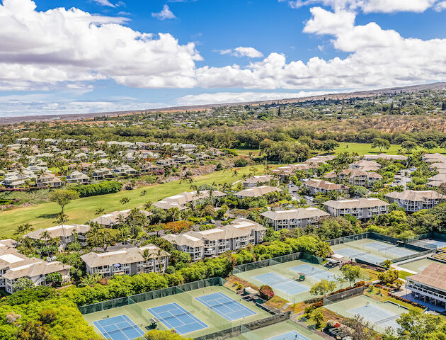 Aerial view of a residential area with multiple buildings, tennis courts, green spaces, and a scenic backdrop under a partly cloudy sky.